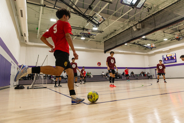 Several young men play futsal in a gym painted white, purple, and gray.