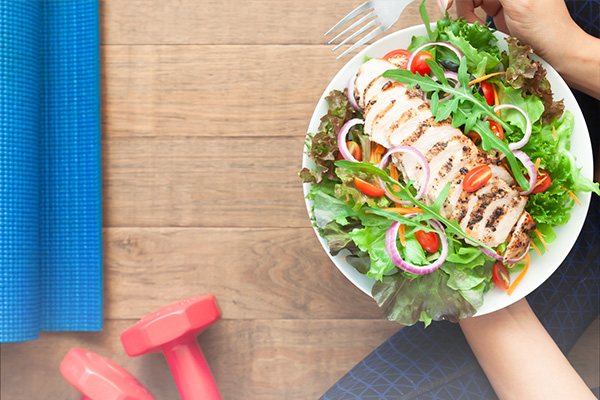 Hands holding a bowl of salad topped with chicken next to a yoga mat and small hand weights.