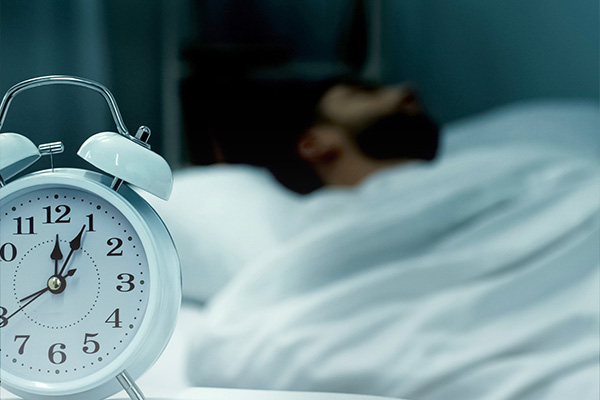 An old-fashioned alarm clock showing the time 12:05, with a blurry, sleeping person in the background.
