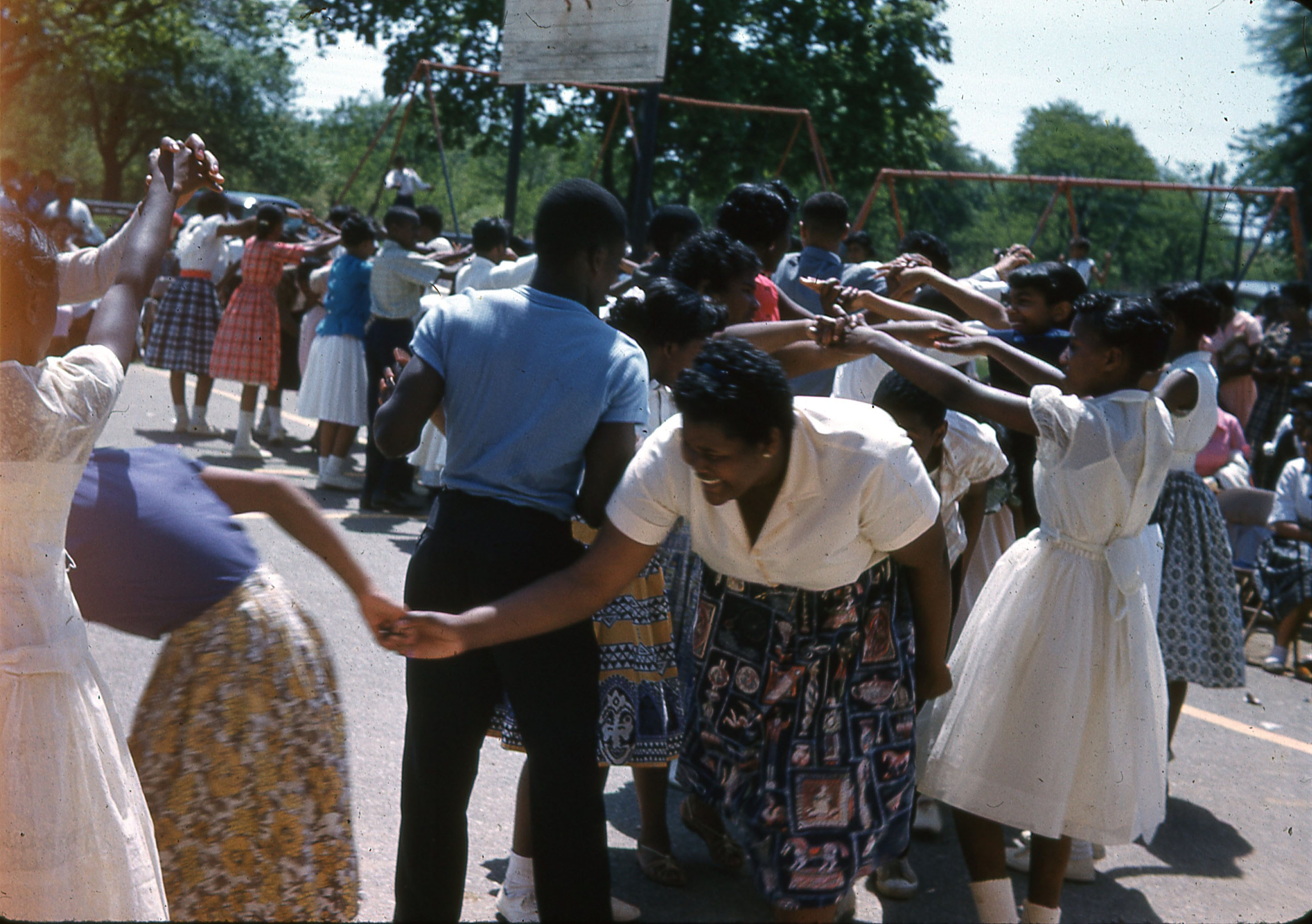 A 1960s candid photograph of group of people participating in a lively outdoor line dance near a playground.