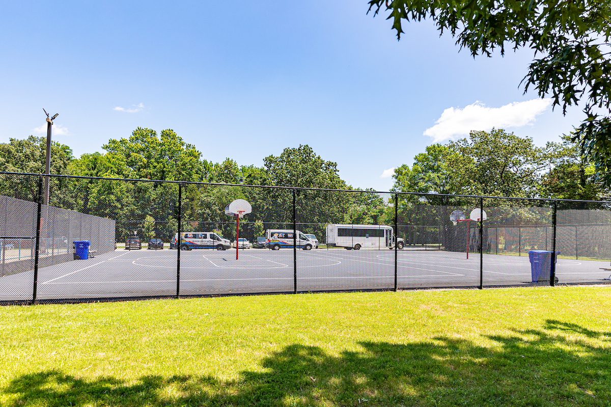 Four sunny, half-court basketball courts in a park, surrounded by a chain-link fence.