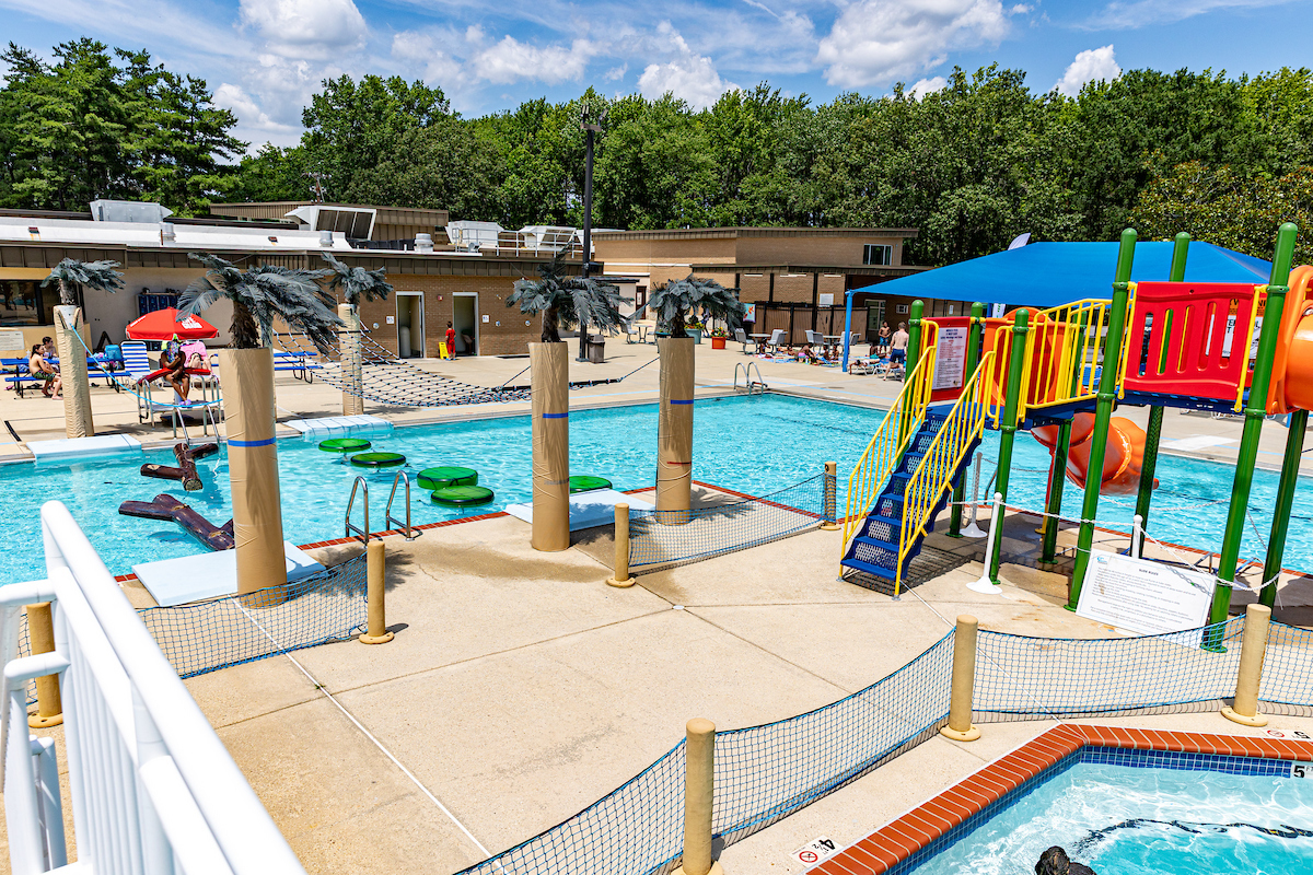 An outdoor splash park with climbing elements, waterslides, and more, next to a large brick building.
