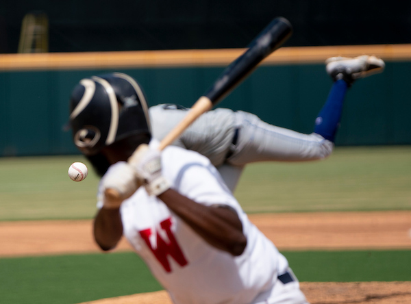 Baseball player swings as pitch nears helmet, bat raised, with blurred field and fence in background.