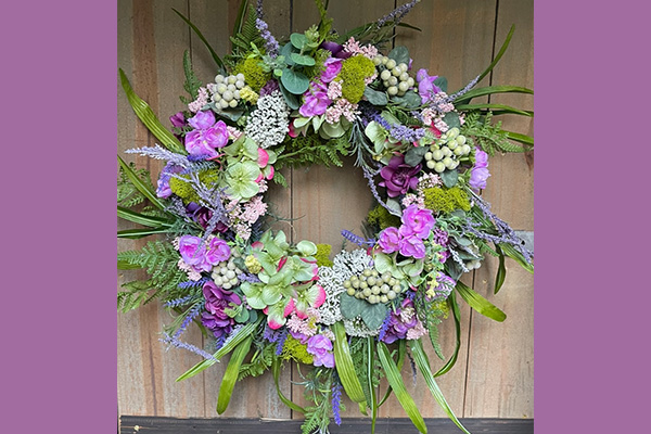 A beautiful wreath composed of various purple flowers, green leaves, moss, ferns, artificial grapes, and more.