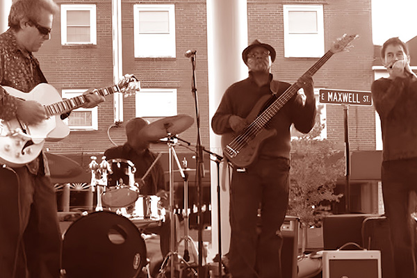 A band performs outdoors near a street sign that says Maxwell St. The band includes a bassist, guitarist, drummer, and singer.