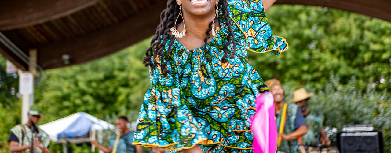 Dancer in a vibrant blue patterned outfit performs onstage while drummers and fellow performers appear in the background.