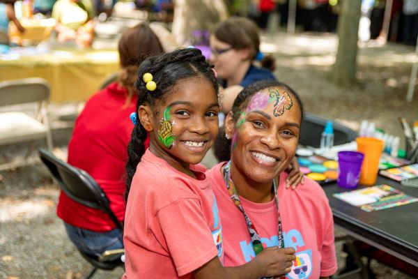 A mother and child with colorful face paint pose, smiling, near a face-painting station in a park.
