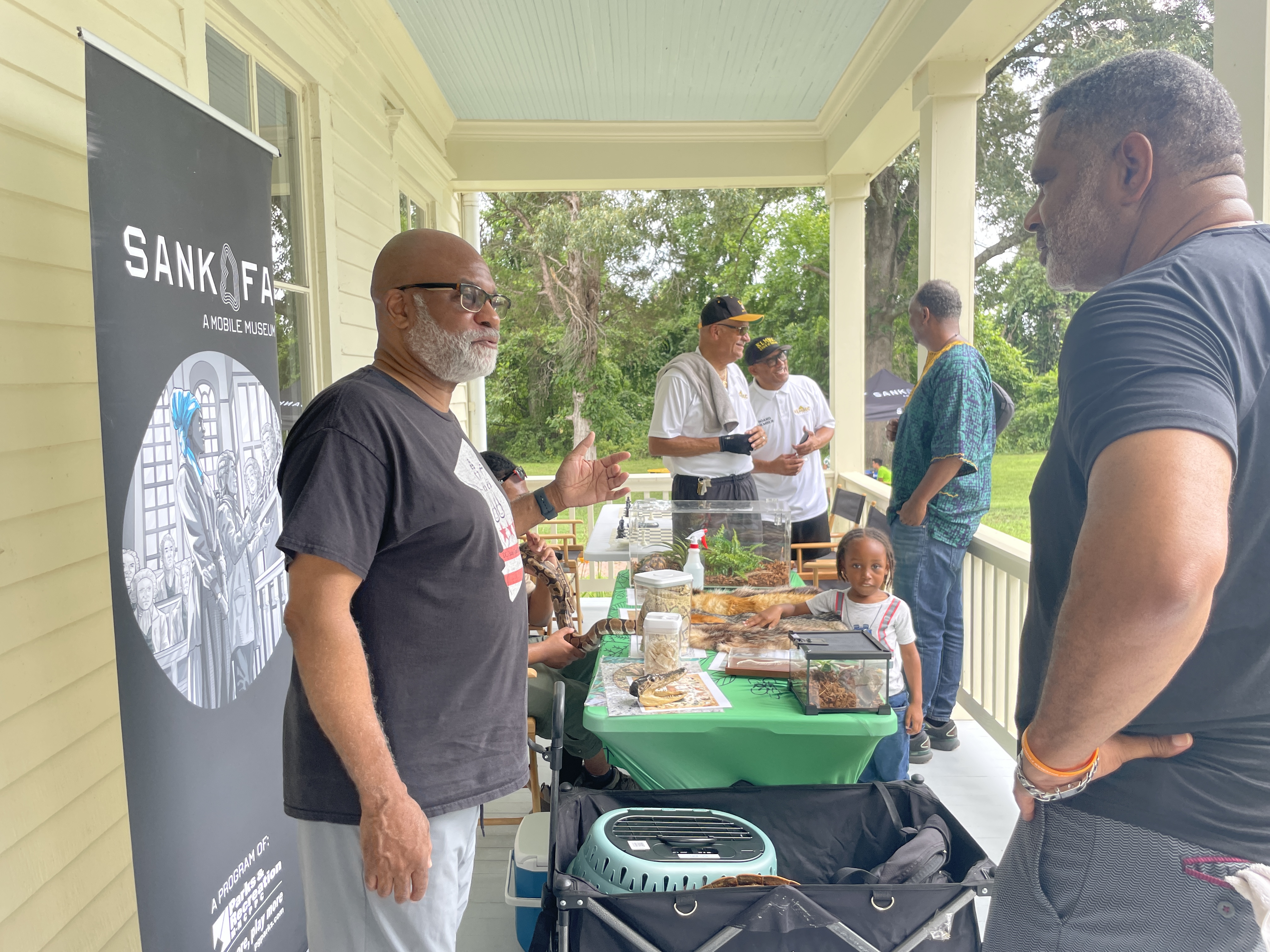 Visitors gather at an educational exhibit on a historic home porch, featuring animal displays and a Sankofa Mobile Museum banner.