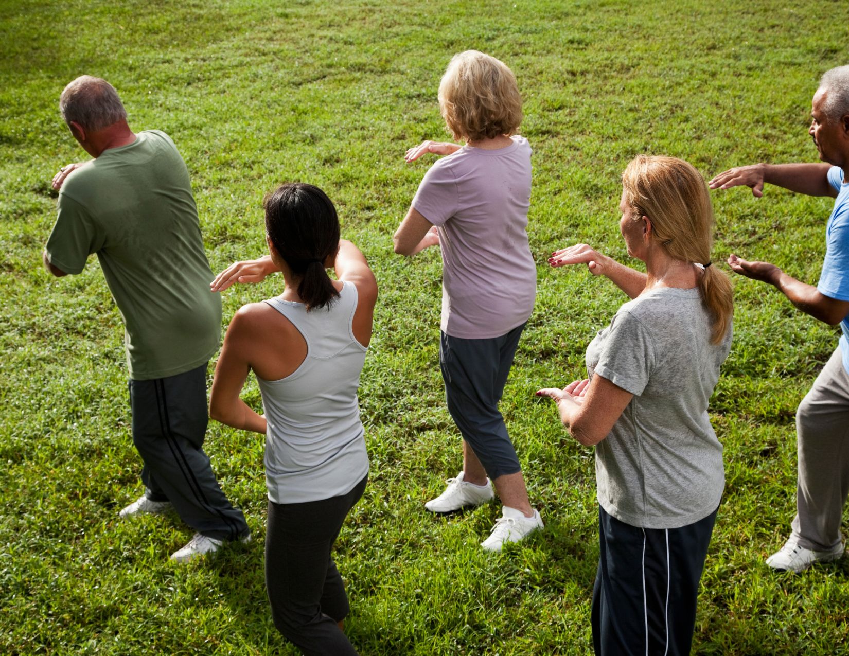 Overhead view of a group practicing fluid Tai Chi arm movements together outdoors on a sunny green lawn.